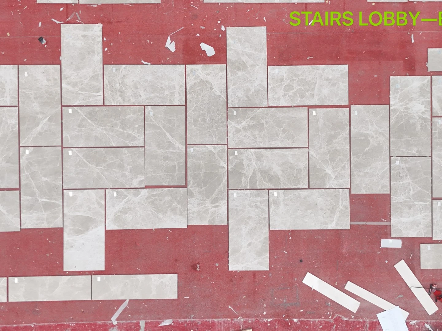 Modern beige marble floor tiles in multiple rectangular sizes arranged on a red factory floor for dry layout inspection, with small identification labels visible on individual pieces. Modern beige marble floor tiles in multiple rectangular sizes arranged on a red factory floor for dry layout inspection, with small identification labels visible on individual pieces.